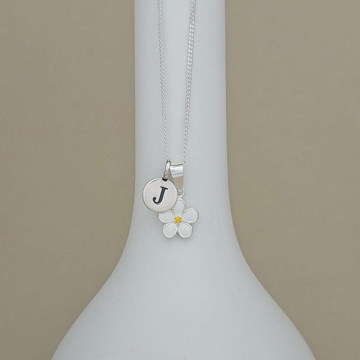 A silver necklace with a round initial 'J' charm and a white flower charm with a yellow center, displayed on a white vase against a beige background.