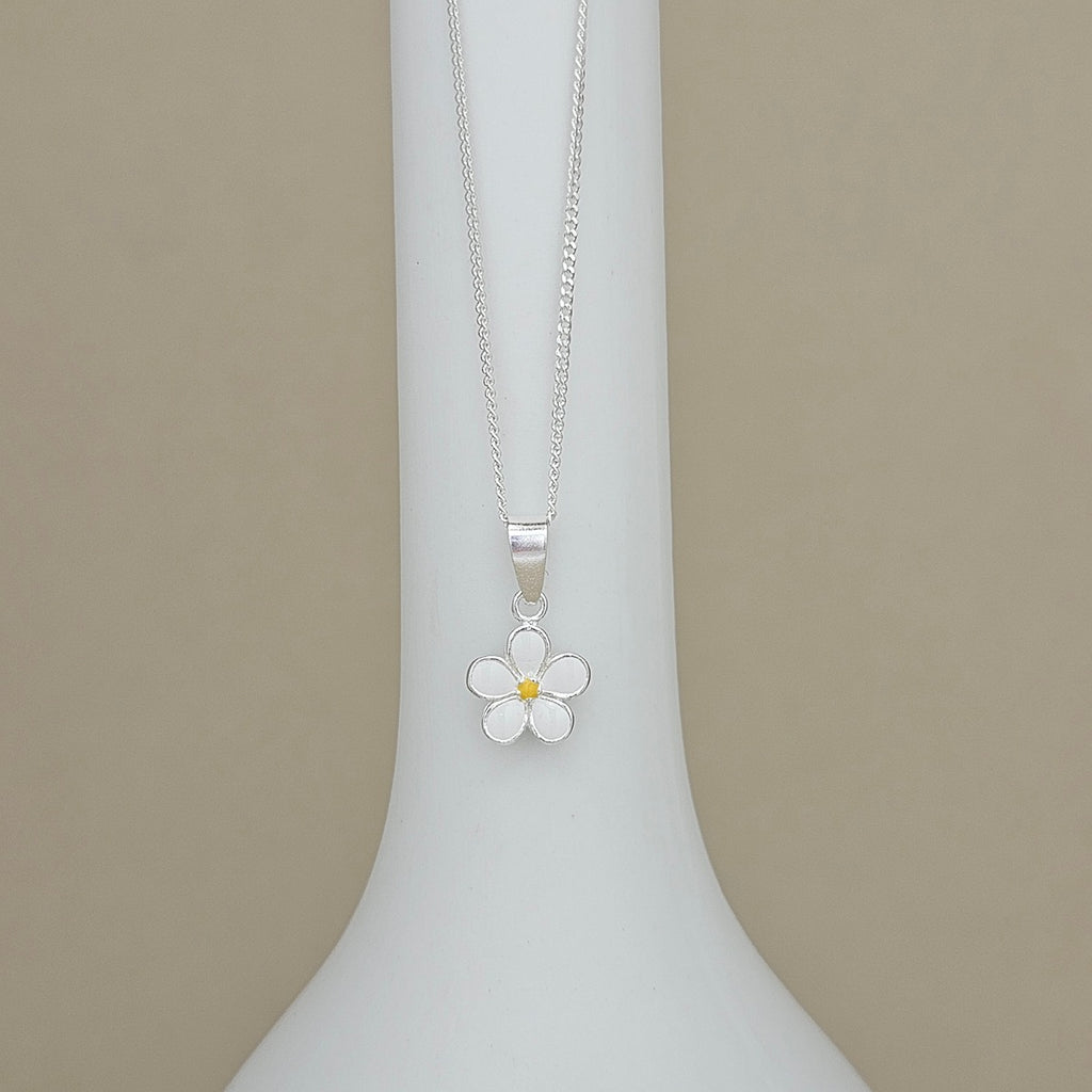A silver daisy flower pendant necklace with a small yellow center, displayed on a white vase against a beige background.