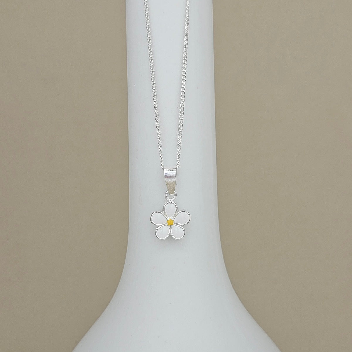 A silver daisy flower pendant necklace with a small yellow center, displayed on a white vase against a beige background.