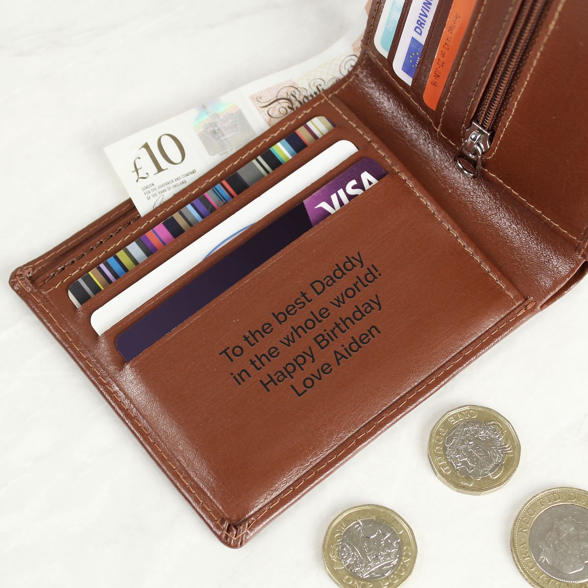 A brown leather bi-fold wallet shown open on a light marble surface, with card slots filled with sample cards, a £10 note, and coins nearby. The wallet has a personalized engraving on the front pocket reading 'To the best Daddy in the whole world! Happy Birthday Love Aiden'.