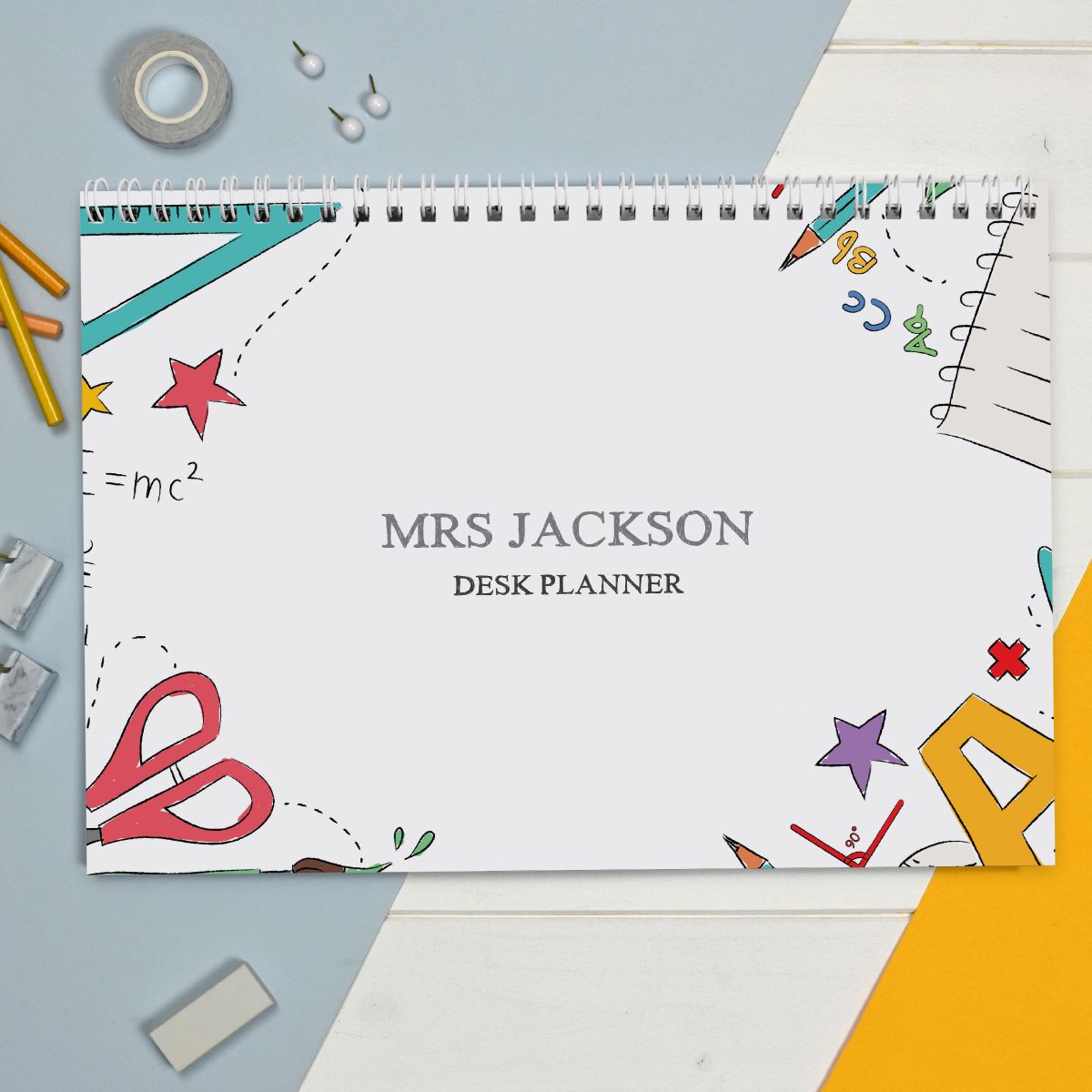 A spiral-bound desk planner titled 'MRS JACKSON DESK PLANNER' lies on a styled flat-lay surface with school and office props including pencils, tape, paper clips, scissors, and geometric paper accents. The planner cover is white with colorful doodles and the text is centered in gray.