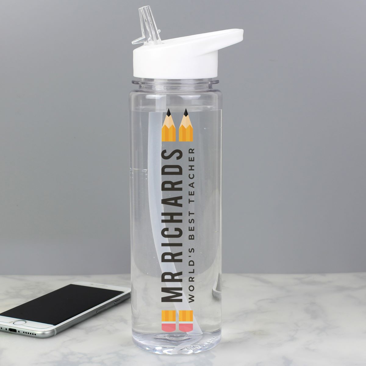 A clear plastic water bottle with a white flip-top lid, featuring a vertical printed design that reads 'MR RICHARDS WORLD'S BEST TEACHER' with two yellow pencils. A smartphone lies on a light marble surface in the background against a gray wall.