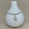 A silver beaded bracelet with a small bear charm and a circular initial 'C' charm, displayed on a white textured glass vase against a neutral beige background.