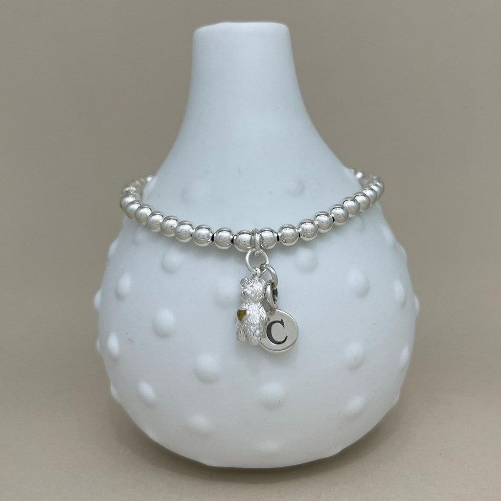 A silver beaded bracelet with a small bear charm and a circular initial 'C' charm, displayed on a white textured glass vase against a neutral beige background.