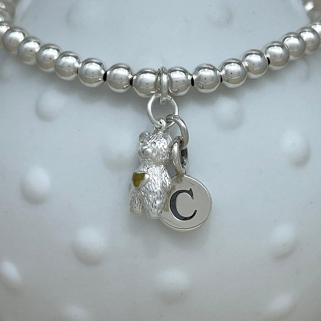 A silver beaded bracelet featuring a textured bear charm and a round initial 'C' disc charm, displayed on a white textured background.
