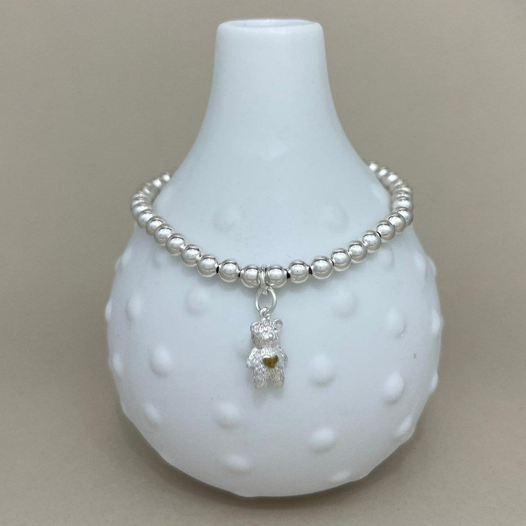 A silver beaded bracelet with a teddy bear charm, displayed on a white textured ceramic vase against a neutral beige background.