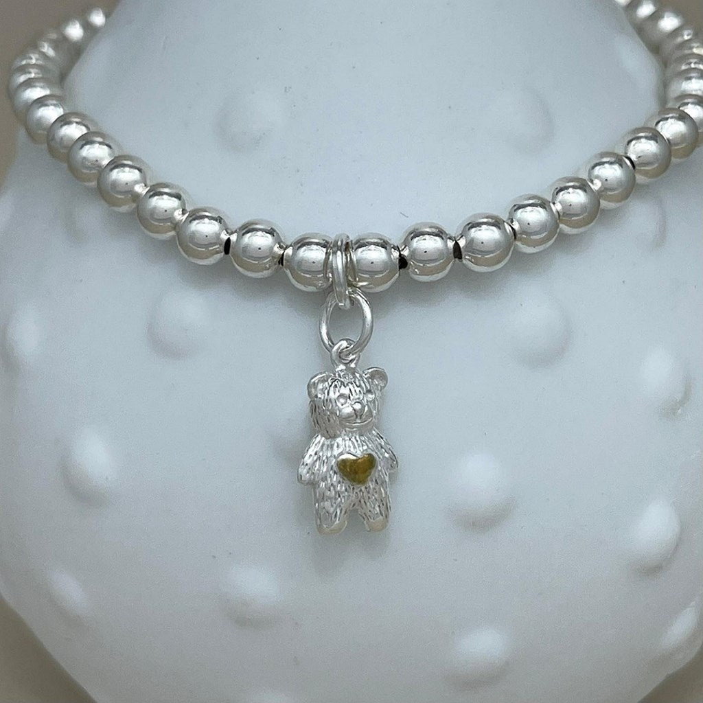 A silver beaded bracelet with a small teddy bear charm with a gold heart, displayed on a white textured bust.