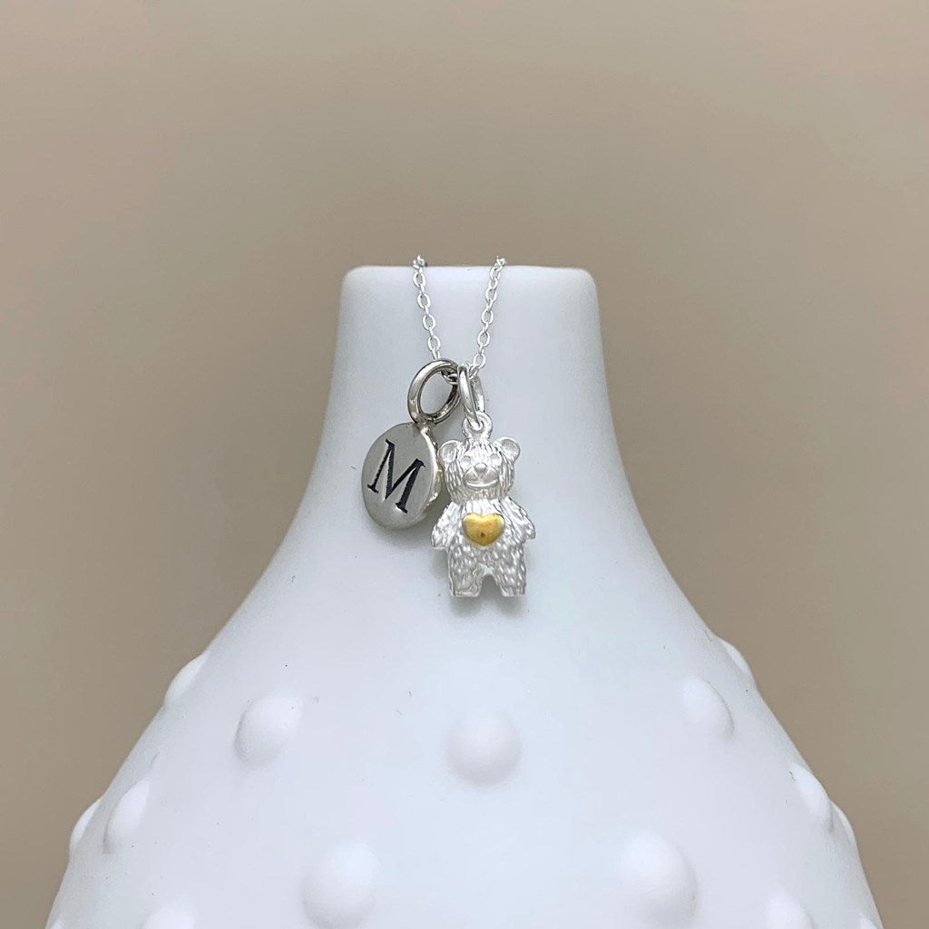 A silver necklace displayed on a white textured vase, featuring a small silver teddy bear charm with a gold heart and a silver oval charm engraved with the letter 'M'.