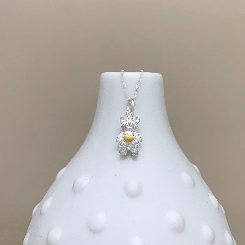 A silver teddy bear pendant necklace with a small gold heart on its chest, displayed on a white textured ceramic vase against a neutral beige background.