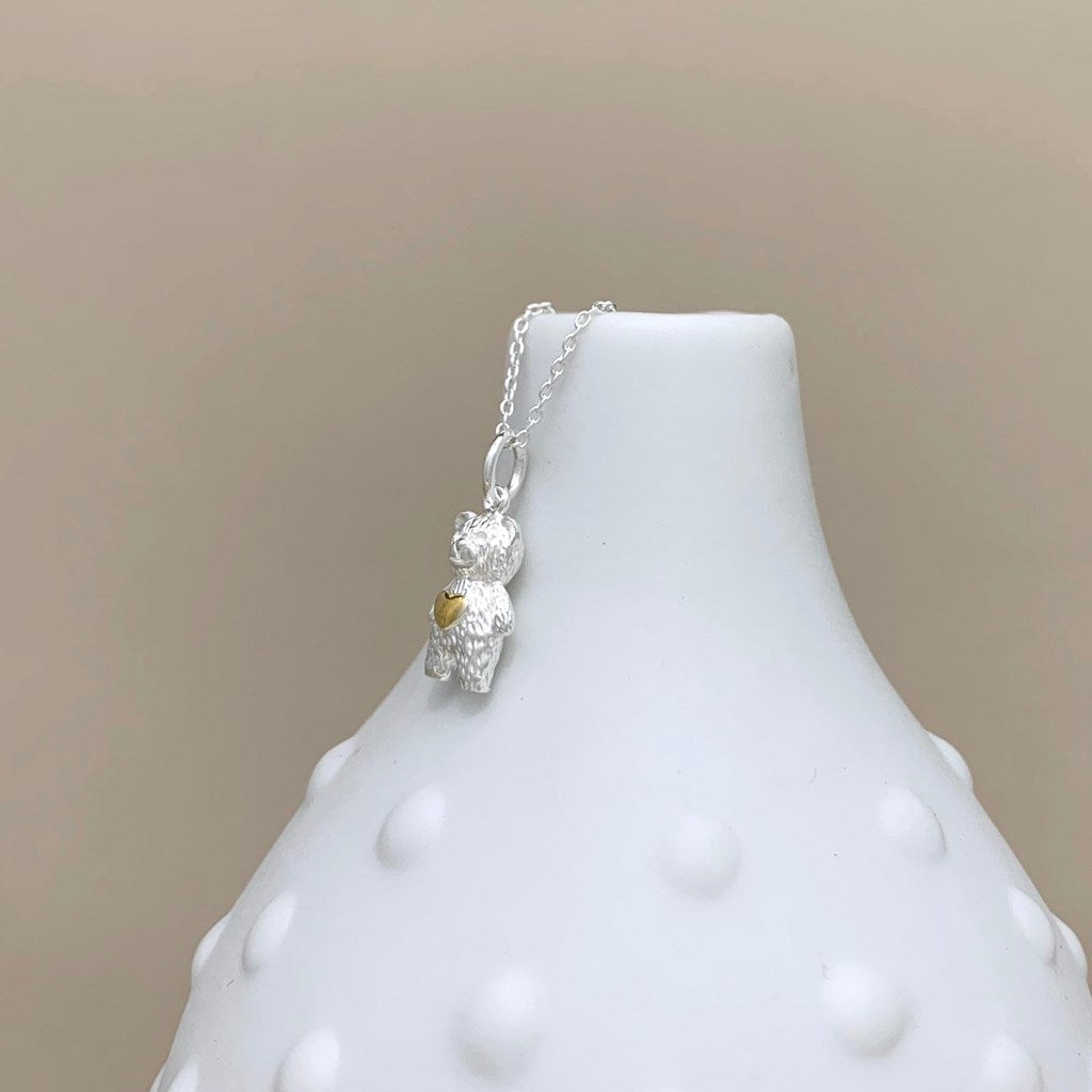A silver necklace with a textured teddy bear pendant and a small gold heart accent, displayed on a white textured ceramic vase against a warm beige background.