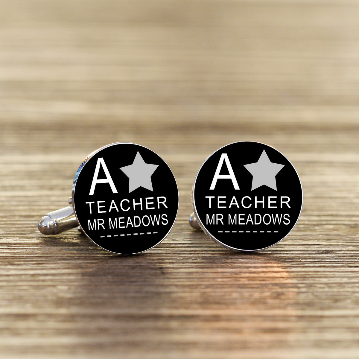 A pair of round black cufflinks with silver-toned metal backs, featuring a white star and the text 'A TEACHER MR MEADOWS' on a wooden surface.