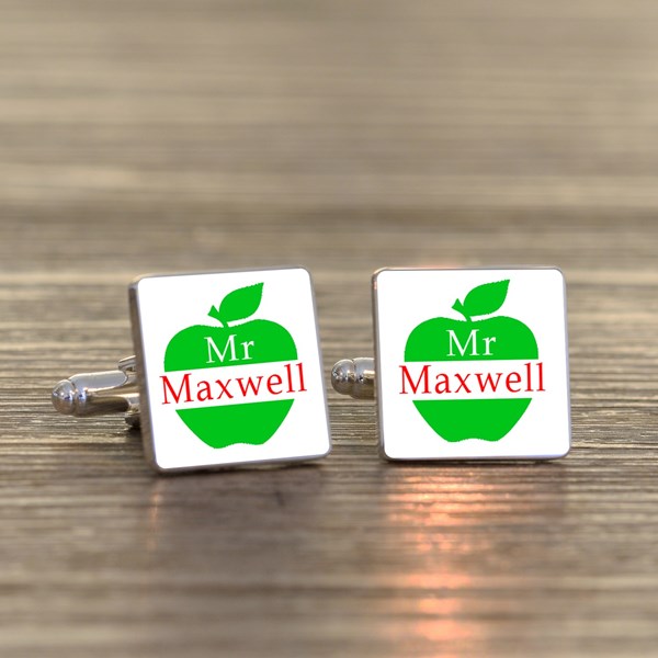A pair of square cufflinks with a white face, featuring a green apple graphic with the text 'Mr Maxwell' in red and white. The cufflinks are displayed on a textured wooden surface.