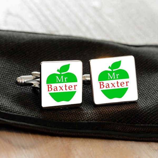 A pair of square cufflinks with a white face, featuring a green apple graphic with the text 'Mr Baxter' in red and white. The cufflinks are displayed on a black textured fabric surface.