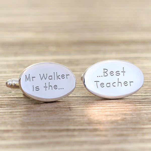 A pair of oval silver cufflinks engraved with personalised names and wording, one reading 'Mr Walker is the....' and the other 'Best Teacher', displayed on a wooden surface.
