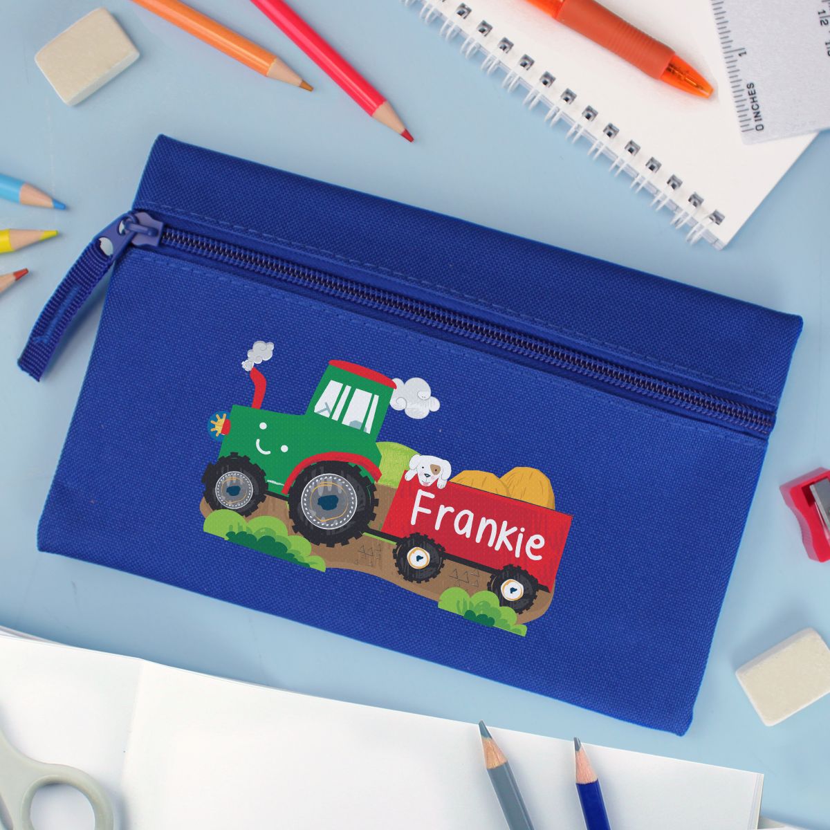 Blue pencil case with a colorful tractor design and 'Frankie' text on a desk with stationery items.