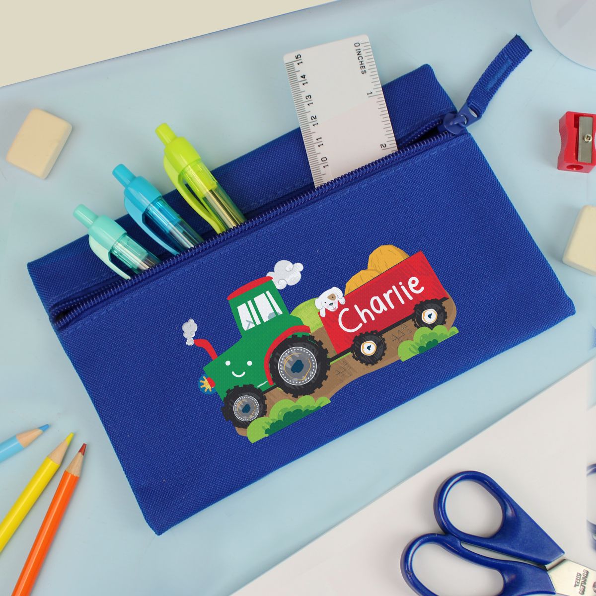 A blue canvas zippered pencil case with a cartoon tractor and trailer design, filled with pens, a ruler, and placed on a light blue desk with school supplies around it.