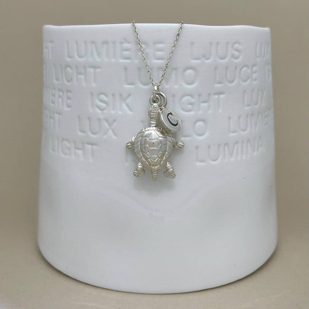 A sterling silver turtle pendant necklace with a letter charm, displayed on a white ceramic candle holder against a neutral beige background.