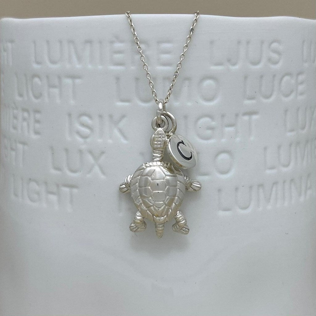A silver turtle pendant necklace with a small circular charm engraved with the letter 'C', displayed on a white ceramic mug with embossed light words.