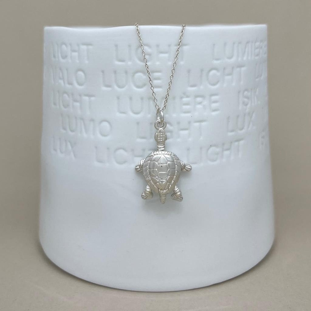 A silver turtle pendant necklace on a delicate chain, displayed against a white ceramic vase set against a neutral beige background.
