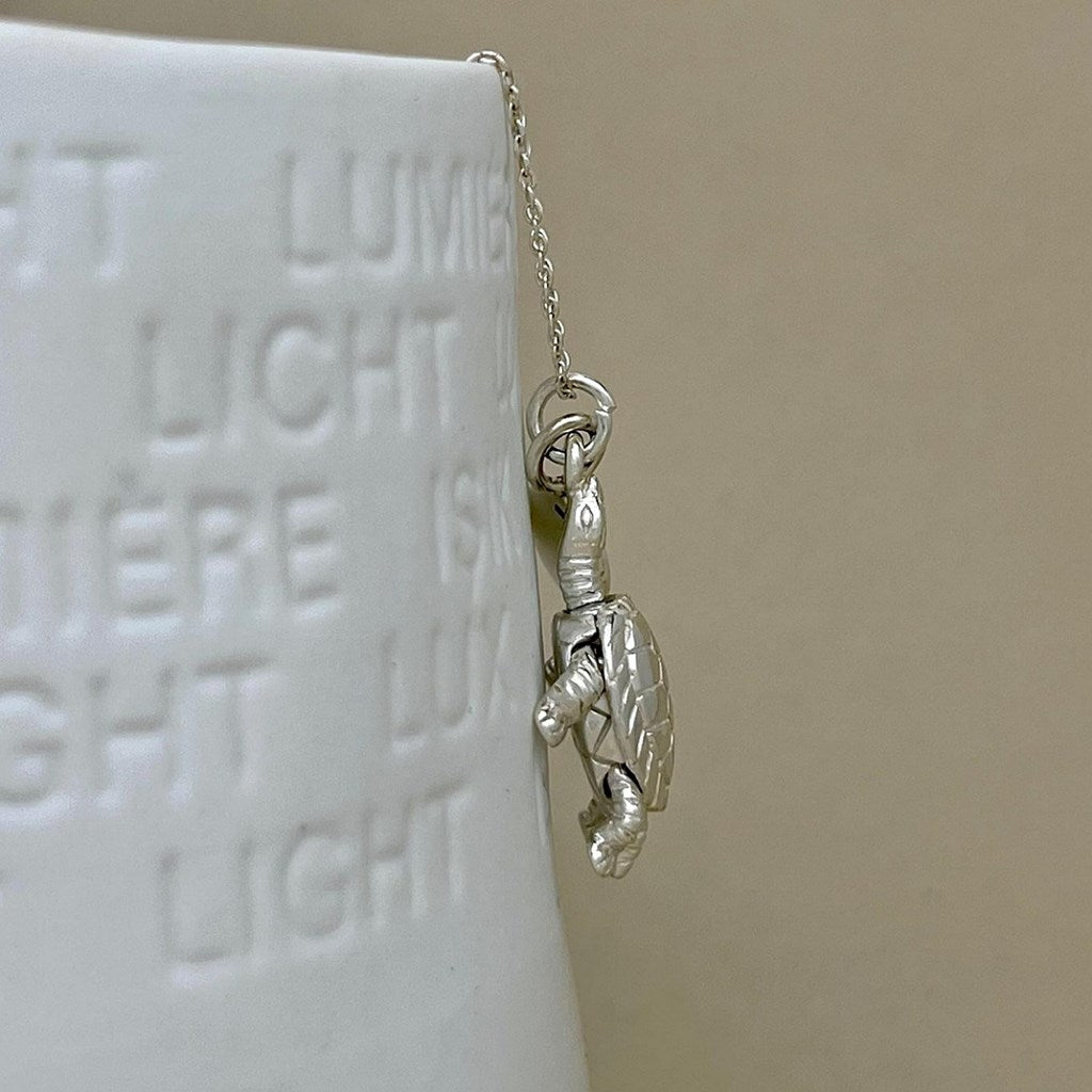 A silver turtle pendant hanging from a delicate chain, positioned next to a white ceramic vase, against a warm beige background.