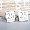A pair of silver square cufflinks on a light grey surface with a blurred rustic wooden background. Both cufflinks show the number 25 and the date 03.02.1996.
