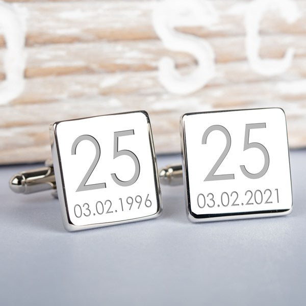 A pair of silver square cufflinks on a light grey surface with a blurred rustic wooden background. Both cufflinks show the number 25 and the date 03.02.1996.