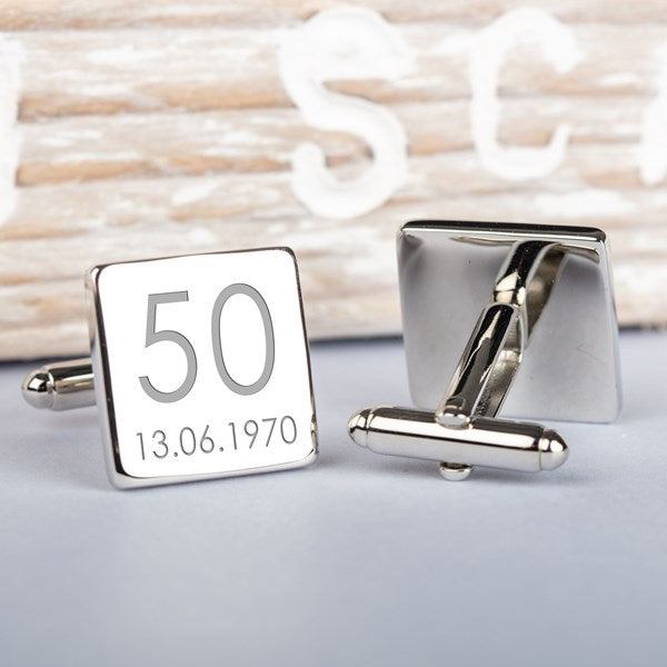 A pair of silver square cufflinks on a light grey surface with a blurred rustic wooden background. One cufflink is shown from the front with the number 50 and the date 13.06.1970.