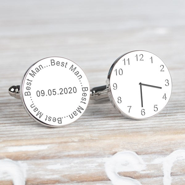 A pair of round silver cufflinks displayed on a light wood background. One cufflink is engraved with the name 'Best Man' and the date, the other cufflink displaying a clock showing a time.