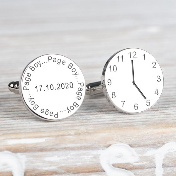 A pair of round silver cufflinks displayed on a light wood background. One cufflink is engraved with the name 'Page Boy' and the date, the other cufflink displaying a clock showing a time.