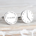 A pair of round silver cufflinks displayed on a light wood background. One cufflink is engraved with the name 'Page Boy' and the date, the other cufflink displaying a clock showing a time.