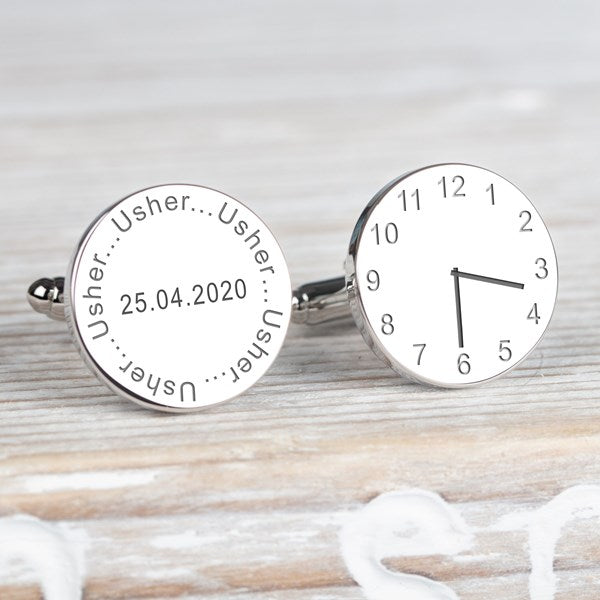 A pair of round silver cufflinks displayed on a light wood background. One cufflink is engraved with the name 'Usher' and the date, the other cufflink displaying a clock showing a time.