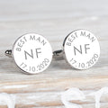 A pair of round silver cufflinks engraved with 'BEST MAN NF 17.10.2020' on a light wood background.