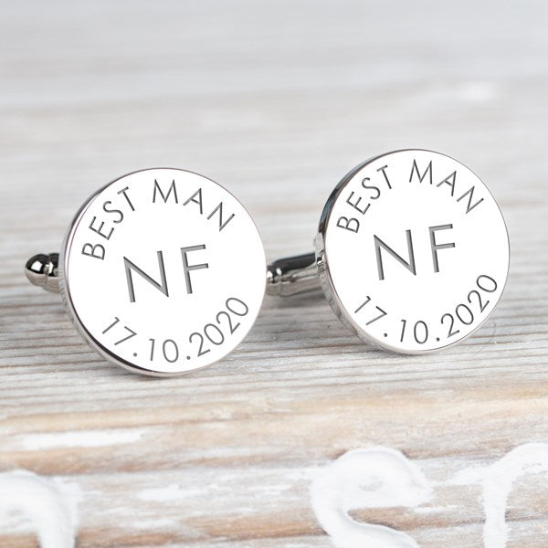 A pair of round silver cufflinks engraved with 'BEST MAN NF 17.10.2020' on a light wood background.