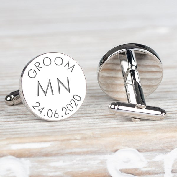 A pair of round silver cufflinks on a light wood surface. One cufflink is shown from the front with engraved text reading 'GROOM', 'MN', and '24.06.2020'.
