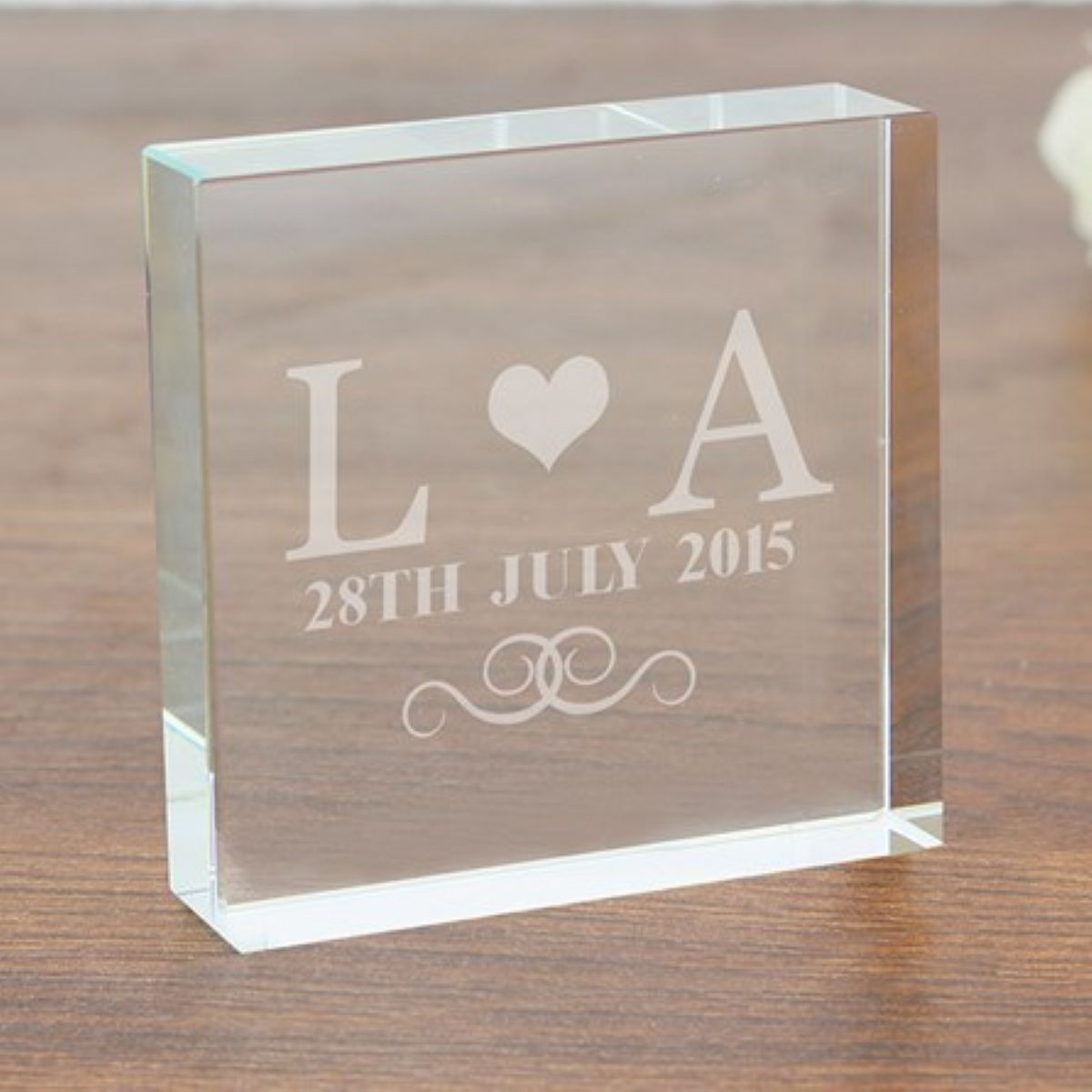 Clear glass block with engraved initials 'L' and 'A', date '28th July 2015', and heart symbol on a wooden surface.
