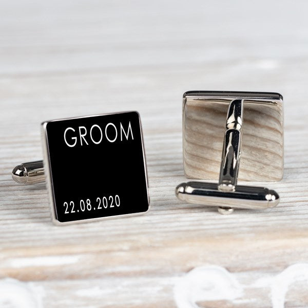 A pair of square silver-toned cufflinks on a light wood surface. One cufflink is shown from the front with a black face engraved with the word 'GROOM' and the date '22.08.2020.