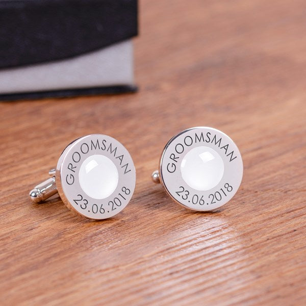 A pair of personalised silver cufflinks with a white circular gem centre and engraved text reading 'GROOMSMAN' and '23.06.2018'.