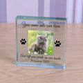 A personalised clear glass block pet memorial plaque standing on a wooden surface. The plaque features a photo of a cat, with black engraved text reading 'You came into our lives' and 'And left paw prints on our hearts Princess 2023', along with two black paw prints.
