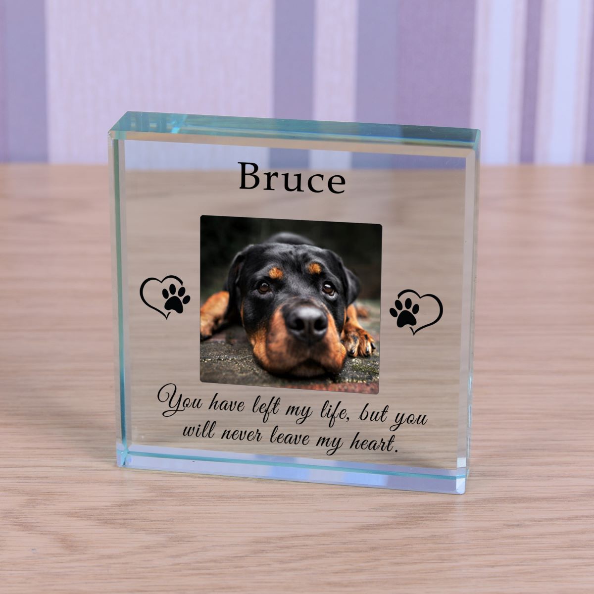A personalised clear glass pet memorial plaque standing on a light wood surface. The plaque features a photo of a Rottweiler dog, the name 'Bruce' at the top, black paw print heart graphics, and the engraved quote: 'You have left my life, but you will never leave my heart.