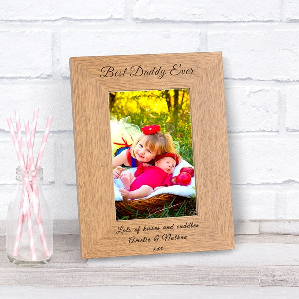 Wooden photo frame with a photo of two children with 'Best Daddy Ever' engraving at the top and 'Lots of kisses and cuddles Amelia and Nathan' at the bottom, displayed against a white brick wall background