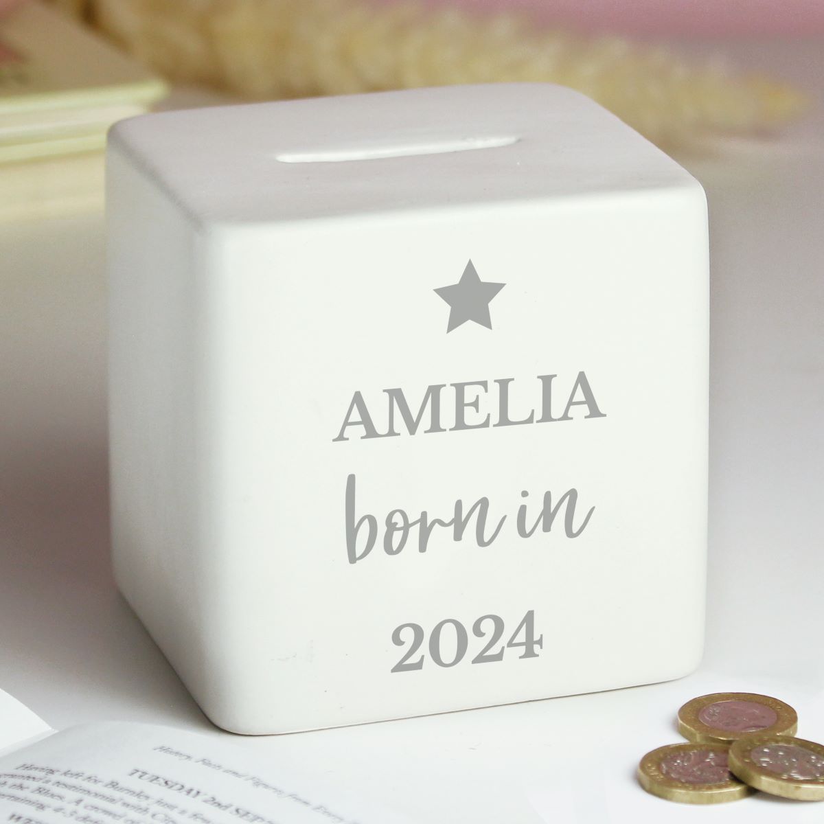A white square ceramic piggy bank with a slot on top, personalized with the name 'AMELIA', 'born in', and the year '2024' in gray lettering with a star above. The piggy bank is styled on a light surface with a book and coins in the foreground.