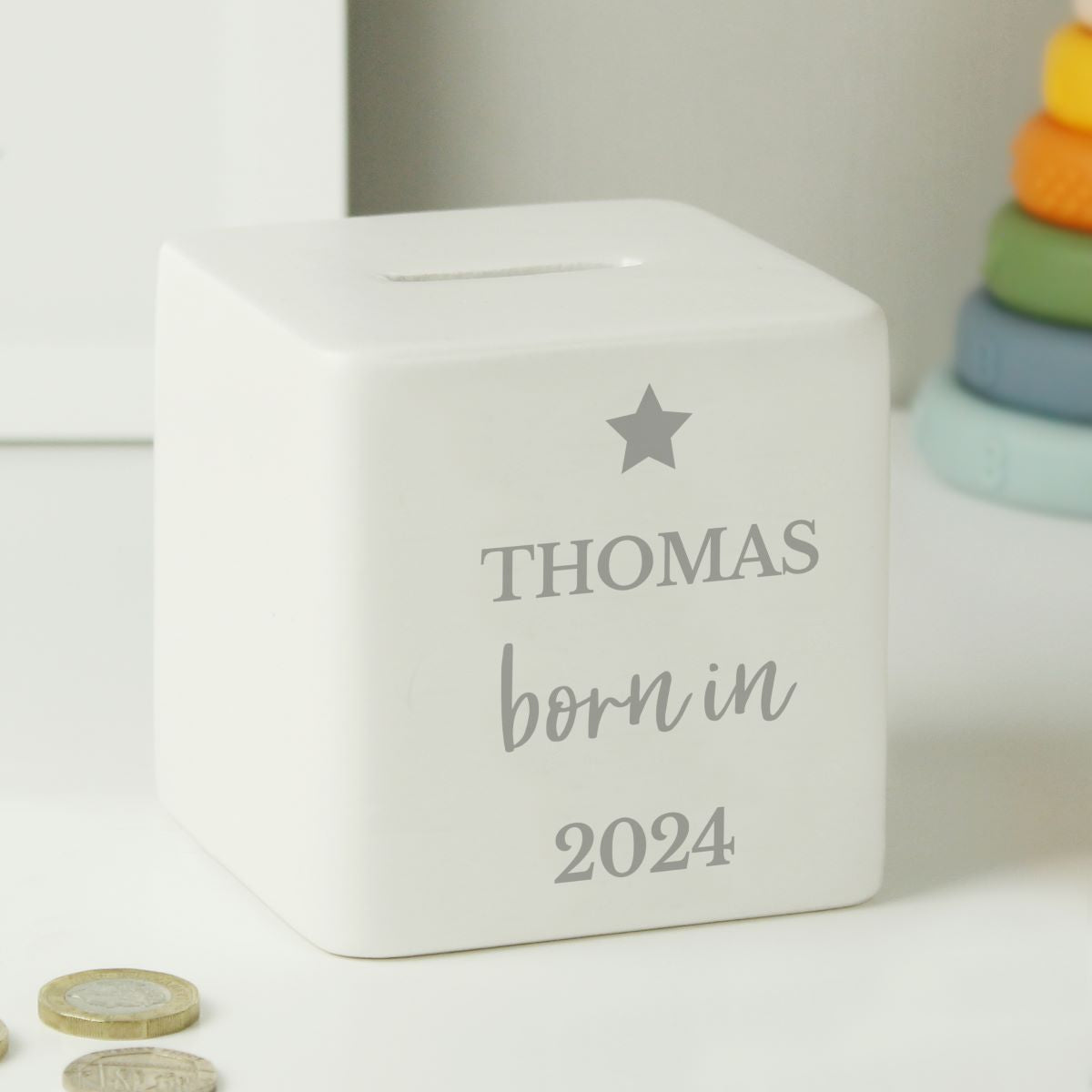 A white square piggy bank with a slot on top, personalized with the name 'THOMAS', 'born in', and '2024' in gray text along with a gray star. The bank is shown on a white surface with coins in the foreground and a colorful toy stack blurred in the background.