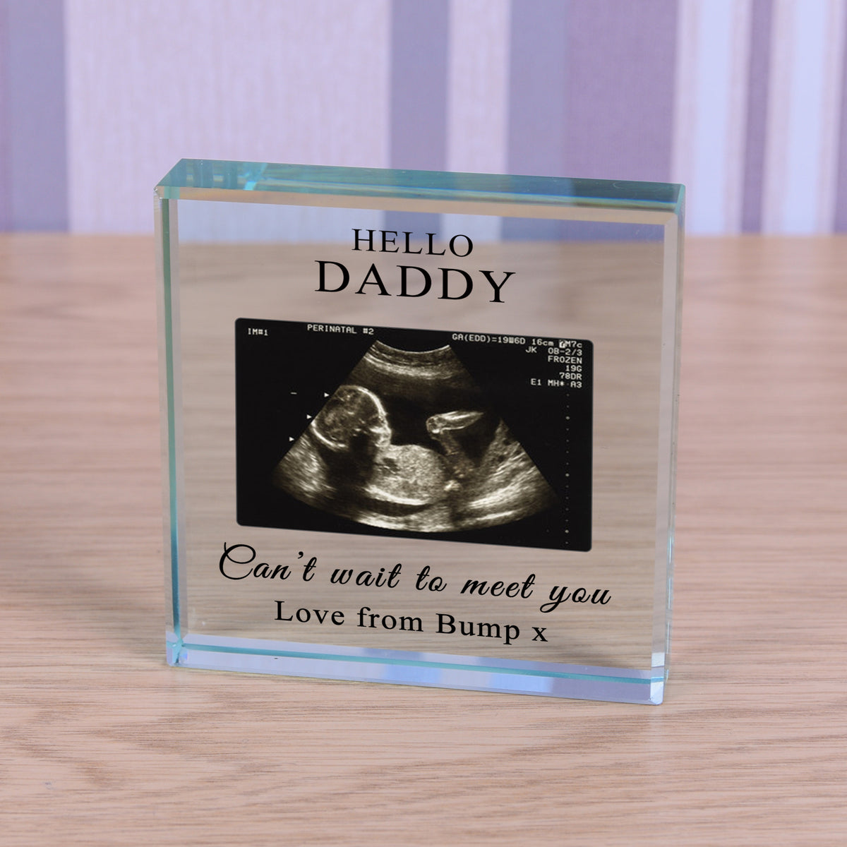 A personalised clear glass block on a light wood surface, featuring a black-and-white ultrasound photo and the text 'HELLO DADDY' and 'Can't wait to meet you Love from Bump x'.