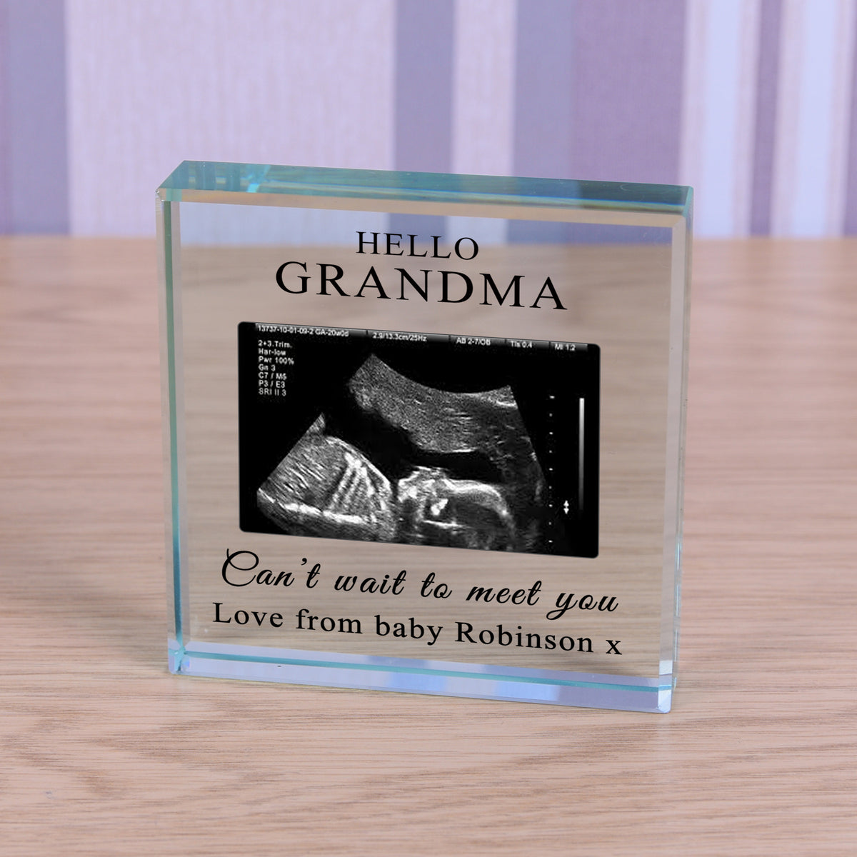 A personalised clear glass block on a light wood surface, featuring a black-and-white ultrasound photo and the text 'HELLO GRANDMA' and 'Can't wait to meet you Love from baby Robinson x'.