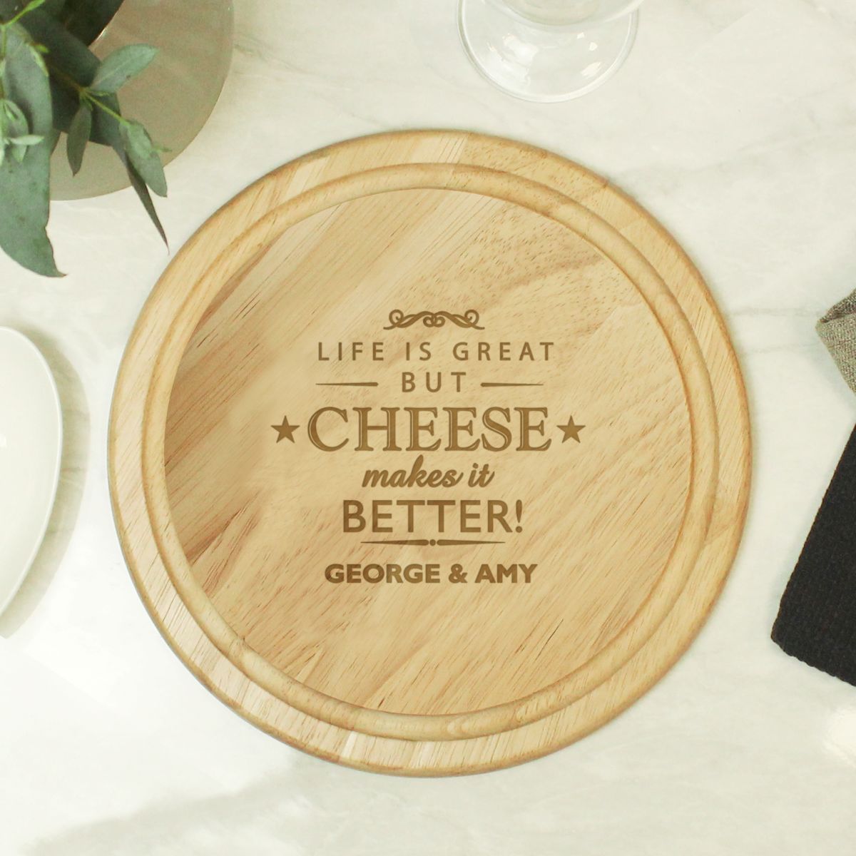A round wooden serving board with engraved text reading 'LIFE IS GREAT BUT CHEESE makes it BETTER! GEORGE & AMY', shown on a light marble-like surface with small decorative props around it.