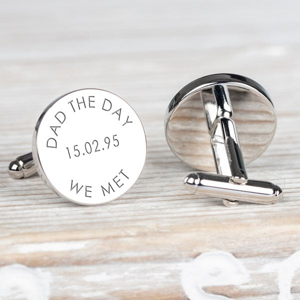 A pair of round silver cufflinks on a light wood surface. One cufflink is shown front-facing with engraved text reading 'DAD THE DAY WE MET' and a date.
