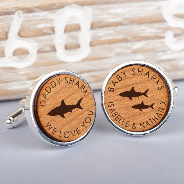 A pair of round silver cufflinks with personalised wooden inserts. One cufflink is engraved with 'DADDY SHARK WE LOVE YOU' and a shark silhouette, while the other is engraved with 'BABY SHARKS ISABELLE & NATHAN X' and two shark silhouettes.