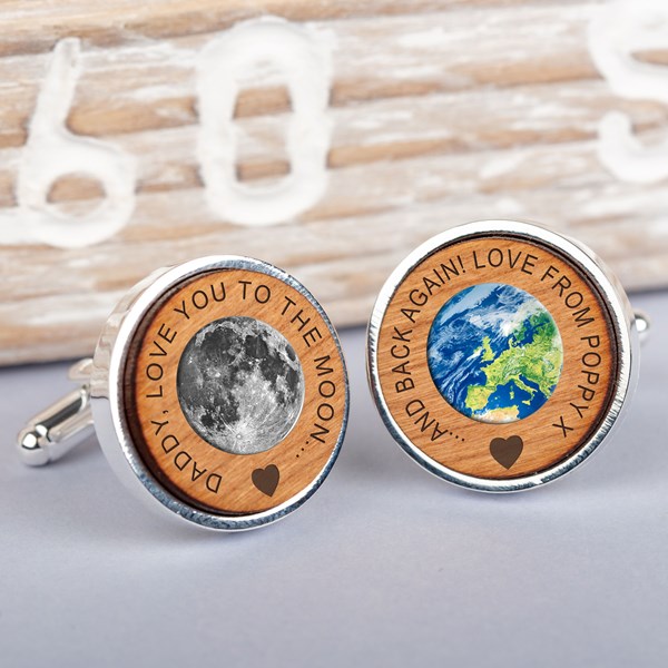 A pair of round silver cufflinks with engraved light brown wood faces displayed on a light grey surface. One cufflink features a detailed engraving of the moon with the text 'Daddy, love you to the moon...'  and the other cufflink features a map of the world with the text '... and back again! love from Poppy.