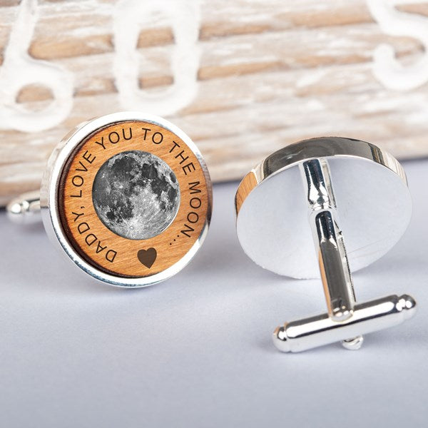A pair of round silver-toned cufflinks displayed on a light grey surface. One cufflink is shown from the front, featuring a circular wooden insert with an engraved moon design and the text 'DADDY I LOVE YOU TO THE MOON.