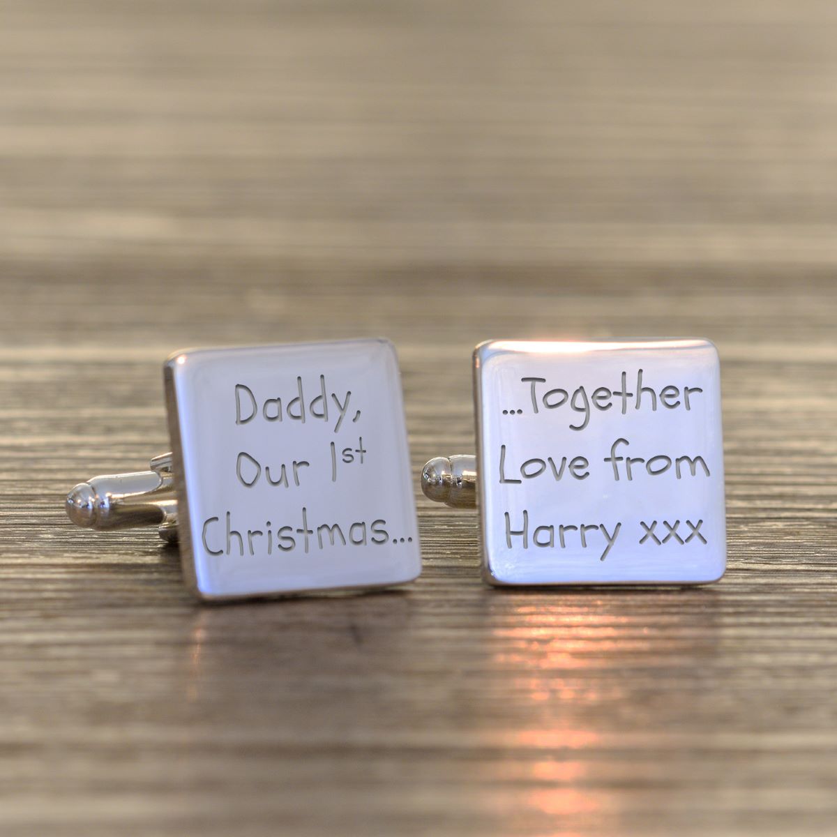 A pair of square silver cufflinks engraved with personalised Christmas messages, resting on a wooden surface. One cufflink is engraved with 'Daddy, Our 1st Christmas and the other '...Together Love from Harry xxx'.